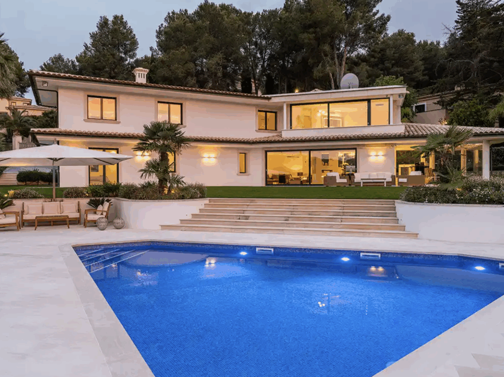 Modern villa at dusk with warmly lit windows; in front of it, a large, blue-lit pool with wide steps and a garden in the background.