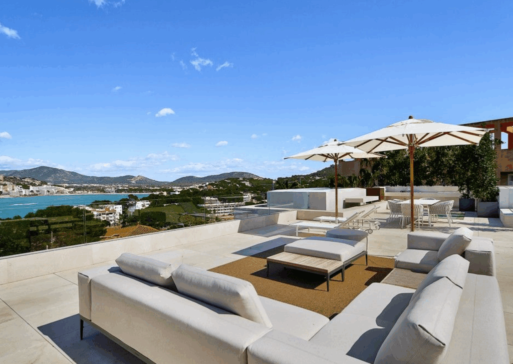 Dachterrasse mit großzügiger Lounge, Sonnenliegen und Sonnenschirmen; Blick über Küste und Meer unter blauem Himmel.