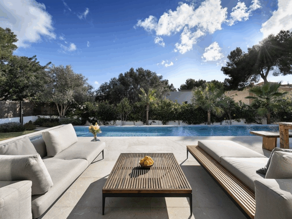 Lounge-Terrasse mit zwei Sofas und Couchtisch vor einem Pool; dahinter Garten mit Bäumen und blauem Himmel mit Wolken.