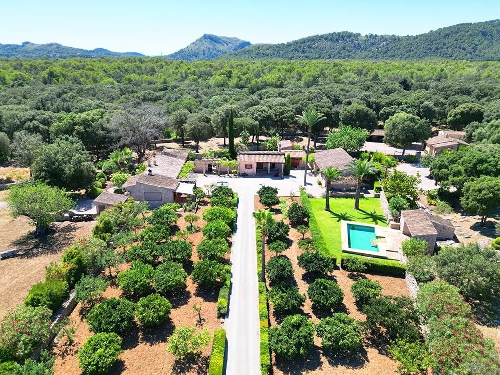 Luftaufnahme einer weitläufigen Wohnanlage mit mehreren Gebäuden, Gartenflächen, Zufahrtsweg und Pool, umgeben von Wald und Hügeln.