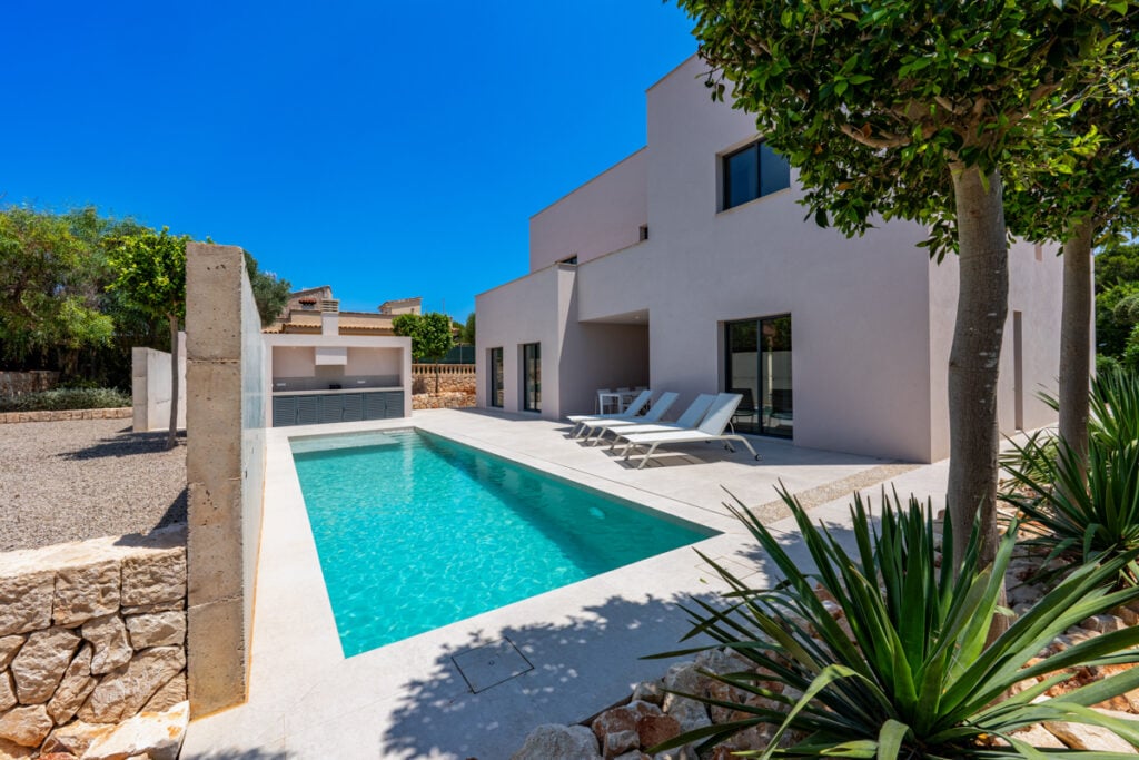 Moderner Außenpool neben heller Villa mit Liegen, umgeben von Bäumen und mediterraner Bepflanzung.