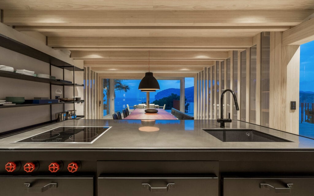 Moderne Küche mit dunkler Kochinsel und offenen Regalen; im Hintergrund große Fensterfront mit Blick auf Pool und Meer bei Dämmerung.