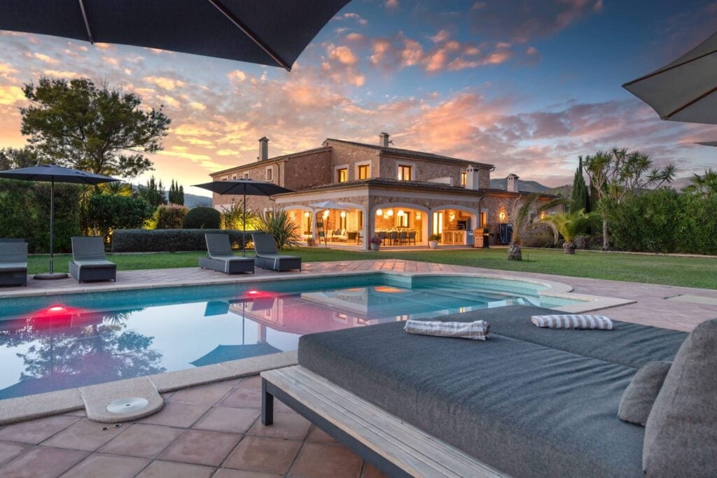 Luxusvilla bei Sonnenuntergang mit beleuchtetem Innenbereich, großer Poolanlage und Lounge-Terrasse im Vordergrund.