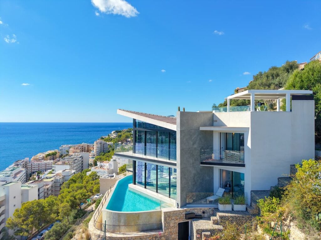 Moderna villa en la ladera con grandes ventanales y piscina infinita; vistas a la ciudad costera y al mar azul bajo un cielo despejado.