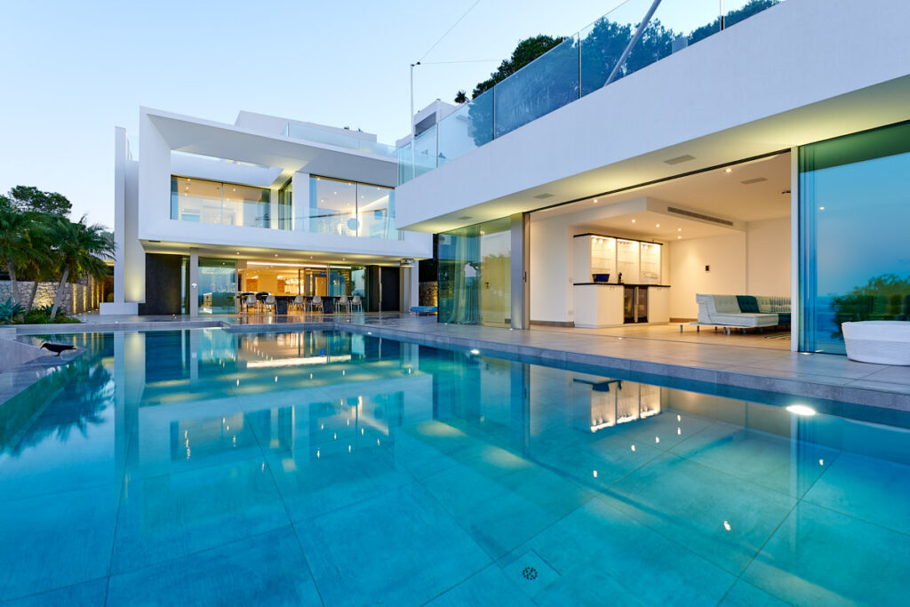Moderne, weiß gestaltete Villa am Abend mit großer Poolanlage; beleuchtete Glasfronten und überdachte Terrasse spiegeln sich im Wasser.