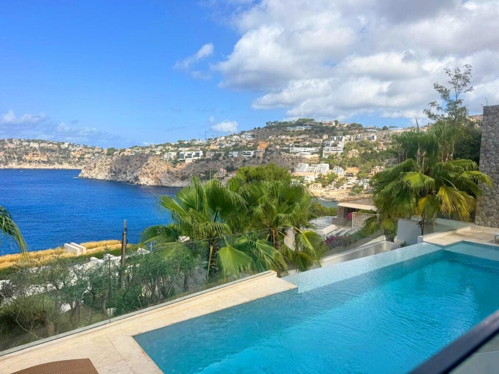 Meerblick von einer Terrasse einer Luxusvilla mit Infinity-Pool und Palmen auf eine felsige Bucht; gegenüber liegen weiße Häuser am Hang unter blauem Himmel.