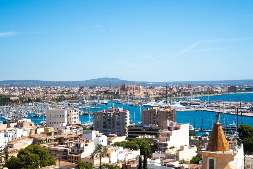 Panoramablick auf einen großen Yachthafen mit vielen Segelbooten; dahinter die Stadt mit markanter Kathedrale und Küstenlinie am Meer.