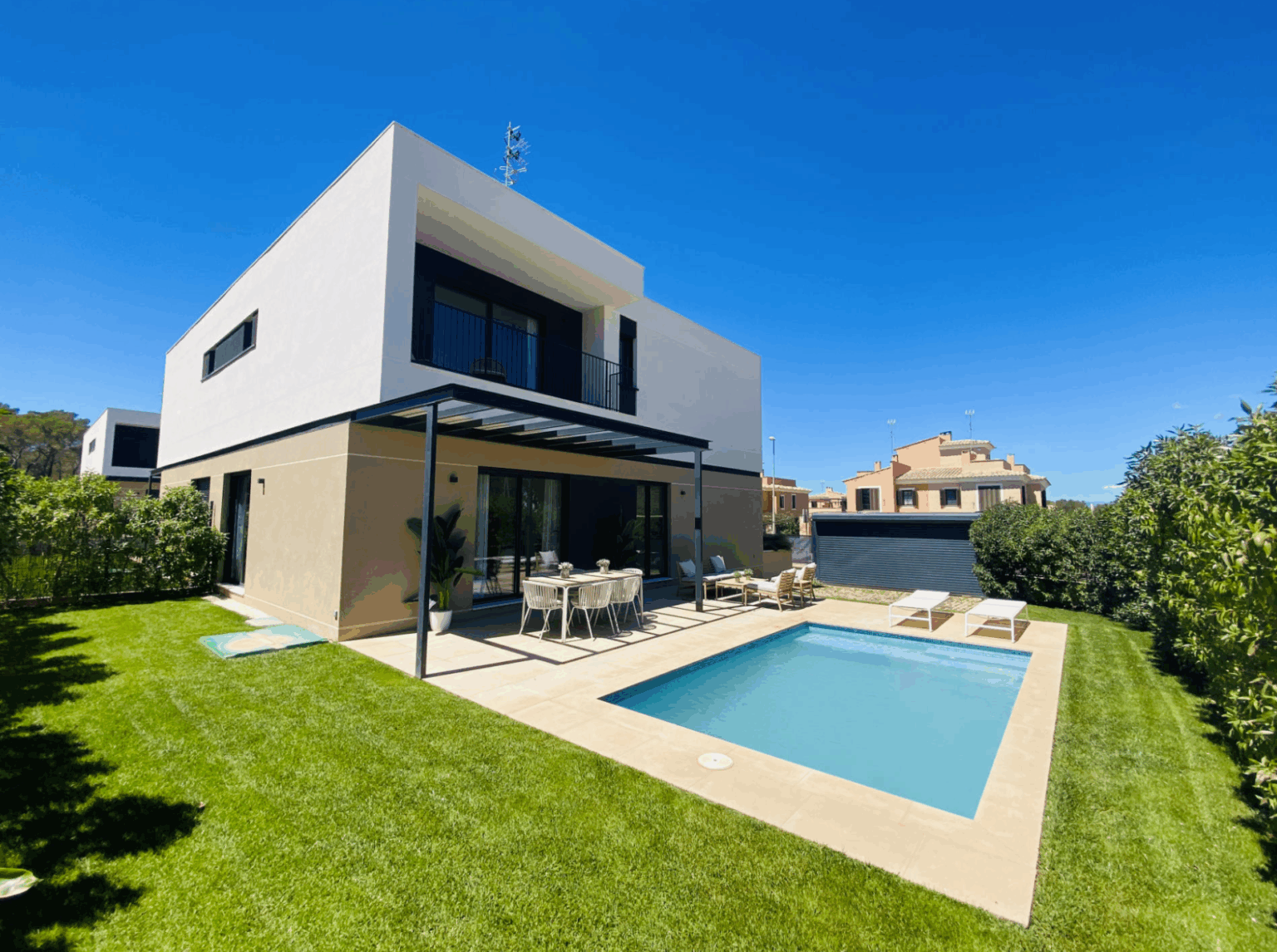 Moderne, zweigeschossige Villa mit Flachdach und überdachter Terrasse; davor ein rechteckiger Pool mit Sonnenliegen auf gepflegtem Rasen unter blauem Himmel.
