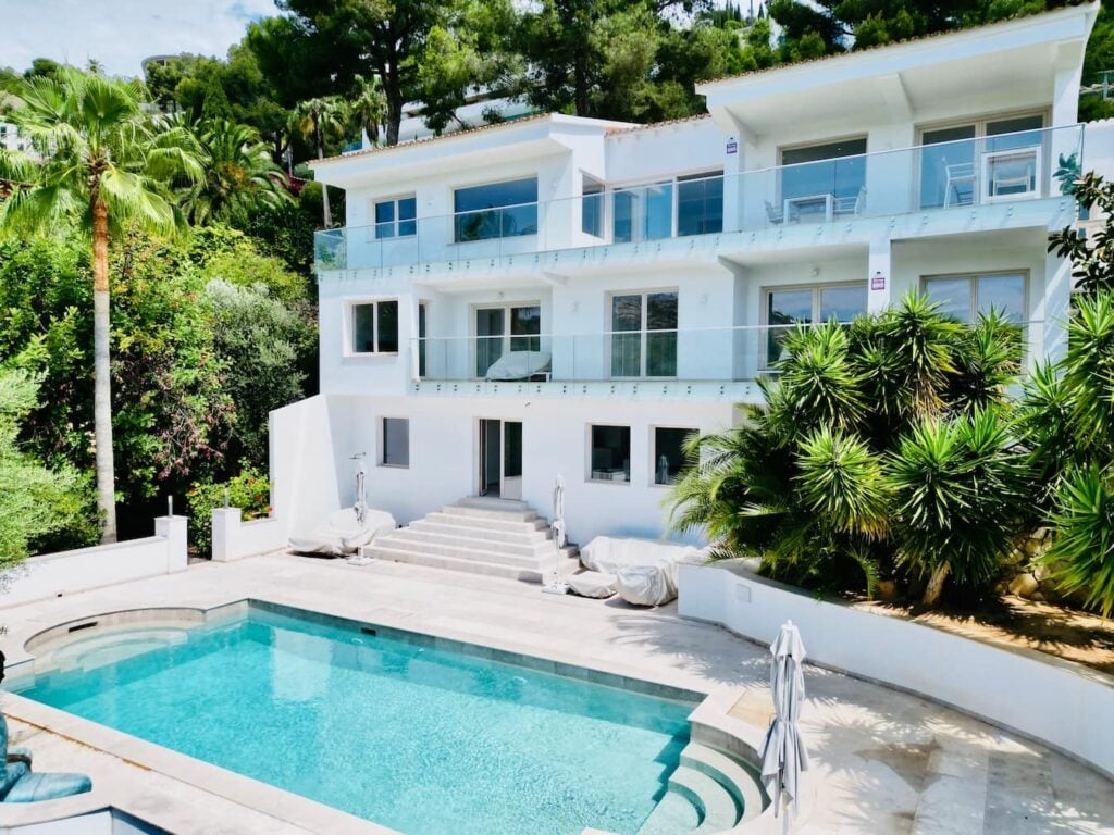 Weiß getünchte Villa mit Glasbalkonen, breitem Treppenaufgang und Pool umgeben von mediterraner Bepflanzung.