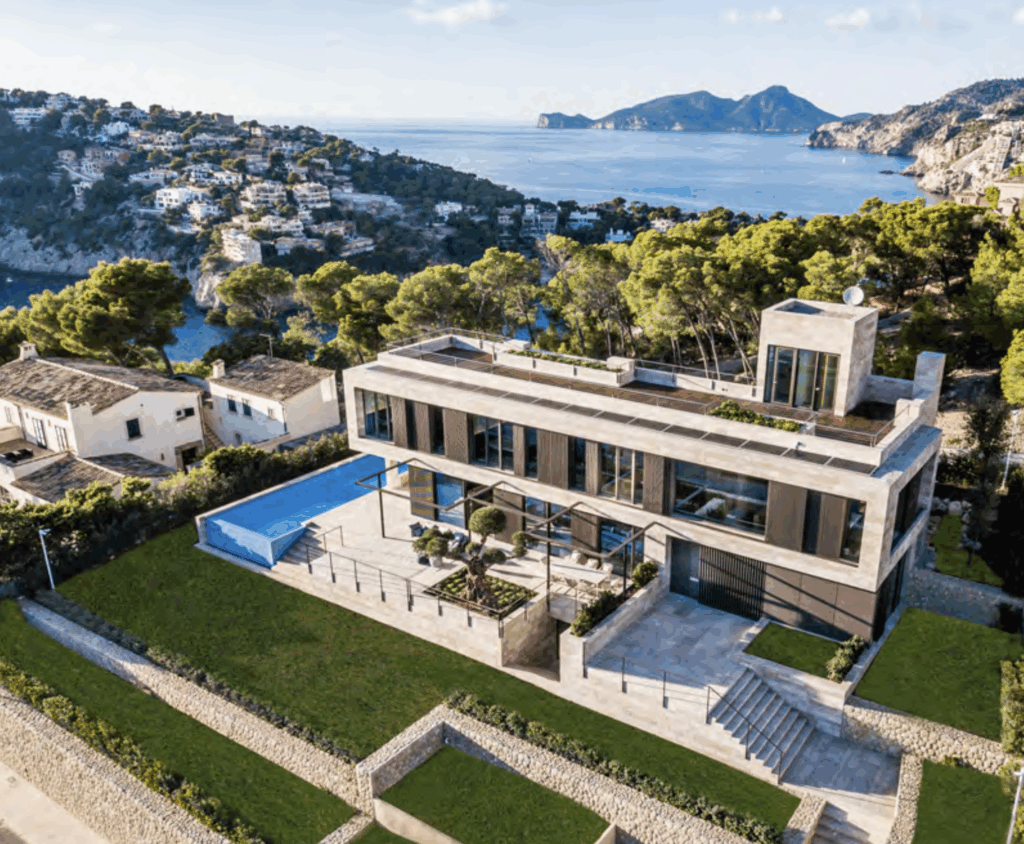 Stone and concrete villa surrounded by a well-kept hedge and stone wall under blue skies, with more stone houses behind it.