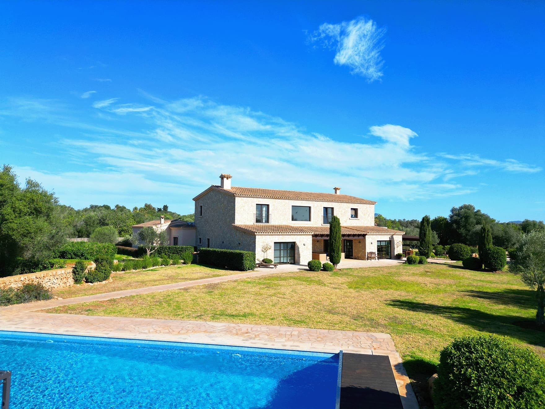 Landhaus mit heller Fassade und Satteldach in weitläufigem Garten; im Vordergrund ein Pool unter blauem Himmel.