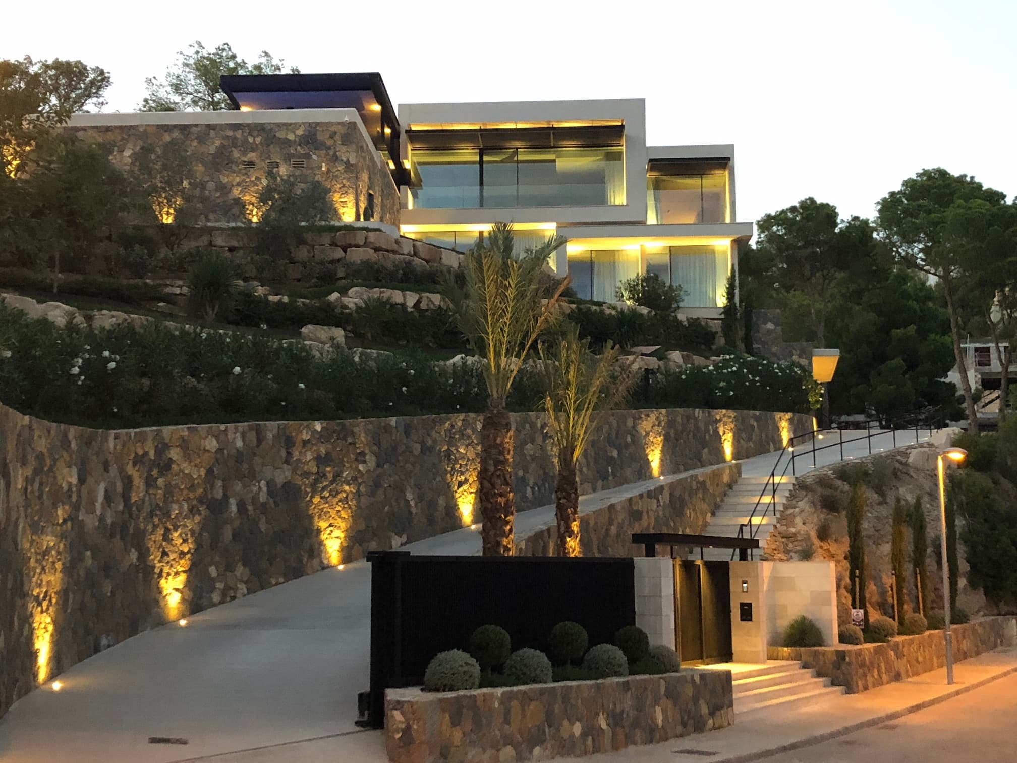 Moderne Luxusvilla am Hang mit Natursteinfassade, großen Glasfronten und stimmungsvoller Beleuchtung in der Dämmerung