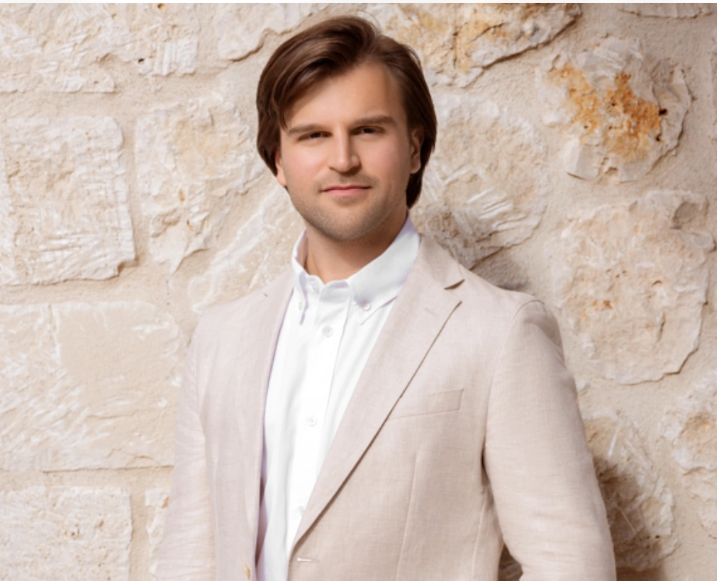 Man in light jacket and white shirt stands in the garden in front of a modern villa with a balcony and round arches.