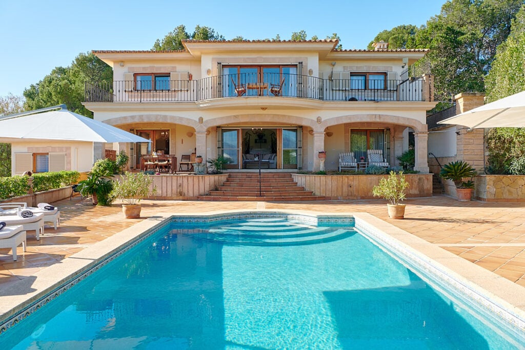 Mediterrane Villa mit zwei Etagen und Balkonen; im Vordergrund ein geschwungener Pool auf sonniger Terrasse mit Sitzbereich.