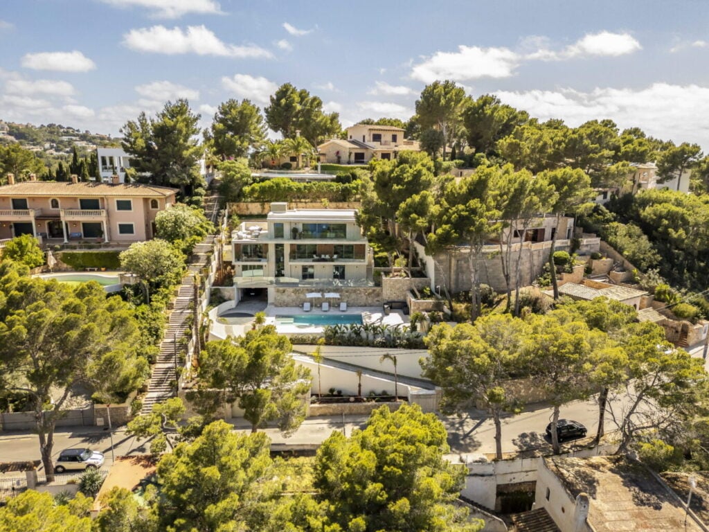 Luftaufnahme einer modernen Villa am Hang mit großer Terrasse und Pool; umgeben von Pinien und Nachbarhäusern in einer ruhigen Wohngegend.